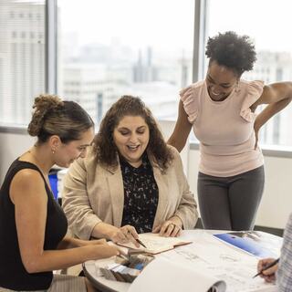Three women collaborate at a table, reviewing documents and sharing ideas in a bright, modern office with city views.