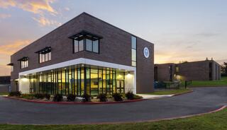 A modern, two-story building with a large glass entrance and brick facade, set against a sunset sky and surrounded by green spaces.