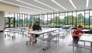A spacious, bright cafeteria features a mother and daughter reading together. A boy sits nearby, engrossed in a book. Large windows offer outdoor views.
