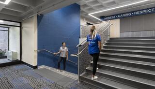 Two women in blue shirts navigate a modern staircase, surrounded by clean lines and motivational words like "Honest," "Caring," and "Respect."