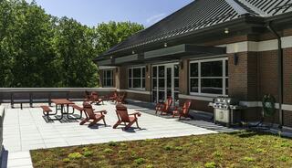 A spacious rooftop patio features red Adirondack chairs, picnic tables, and a grill, surrounded by lush greenery and a clear blue sky.