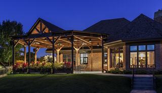 A modern house features a spacious covered patio with wooden beams, surrounded by colorful landscaping, illuminated in soft evening light.