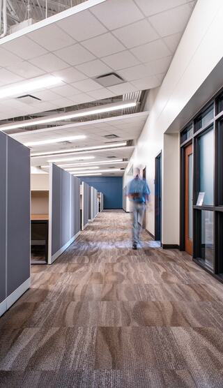 A modern office corridor lined with cubicles, featuring neutral tones and a blurred figure walking towards the camera.