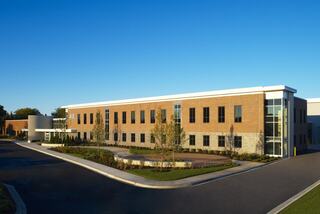 A modern, two-story brick building features large windows and landscaped surroundings, set against a clear blue sky.