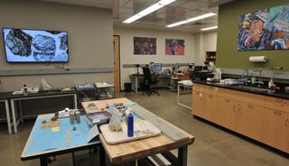 A modern laboratory features workstations with equipment, samples, and monitors displaying rock images, along with colorful artwork on the walls.