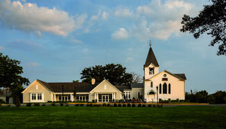 A charming white church with a tall steeple, accompanied by a modern building, set against a backdrop of blue skies and greenery.