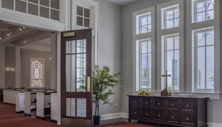 A welcoming church entrance features large windows, a wooden door, a dark cabinet, a potted plant, and sunlight illuminating the interior.