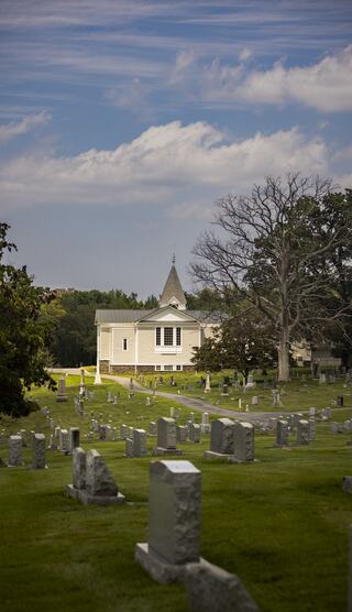 A serene cemetery scene features scattered gravestones in lush green grass, with a quaint church in the background under a cloudy sky.