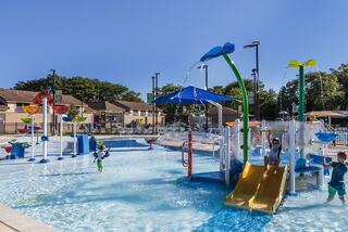 A vibrant water playground features colorful slides, fountains, and kids enjoying splashes on a sunny day, surrounded by green trees and houses.
