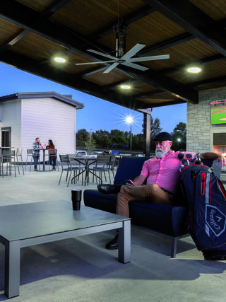 A relaxed man in a pink shirt sits on a couch with a golf bag nearby, while others gather in the background under evening lights.