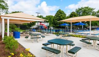 Outdoor pool area featuring shaded picnic tables, a vibrant water slide, and lounge chairs surrounded by greenery under a blue sky.