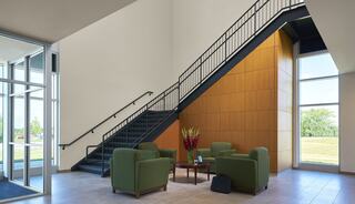 A modern lobby features a sleek staircase, green seating area, and large windows, creating an inviting and bright space.