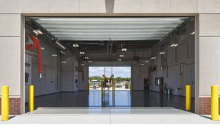 A spacious garage with a large open bay door, showcasing bright lighting and minimal equipment, lined with yellow safety bollards.