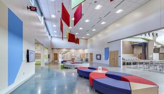 A modern, bright interior features colorful seating and hanging decorations, creating an inviting communal space. Large windows enhance the light.