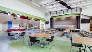 A spacious cafeteria features movable round tables, a stage with curtains, vibrant wall colors, and a modern design.