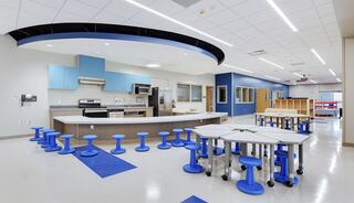A modern kitchen space features a central island, blue accents, multiple cooking appliances, and colorful stools around hexagonal tables.