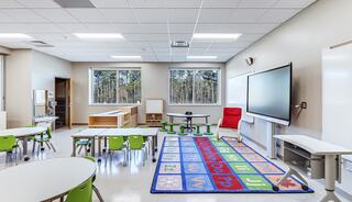 A bright classroom features circular tables, green chairs, a colorful alphabet rug, a large screen, and windows with a view of trees.