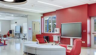 A modern, vibrant classroom features red accent walls, flexible seating, and large windows, creating an inviting learning environment.