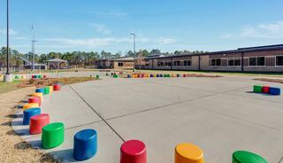 A spacious outdoor area features colorful, cylindrical seating arranged in a circle, surrounded by a grassy landscape and modern school buildings.