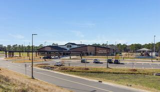A modern school building with a spacious parking lot and a playground, surrounded by grassy areas and trees under a clear blue sky.