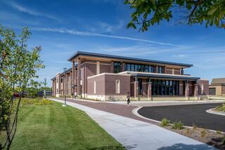 A modern, brick building with large windows surrounded by greenery and a paved walkway, under a clear blue sky.