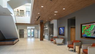 A modern lobby featuring natural light, wooden accents, and seating areas. Two screens display information, enhancing the welcoming atmosphere.