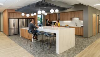 A modern kitchen with a large island, stylish stools, wooden cabinets, and friendly conversation among three people in a bright space.