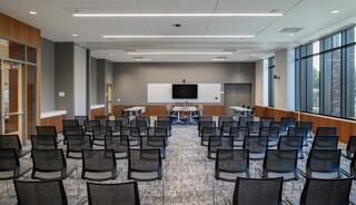 A modern conference room featuring rows of black chairs, a central presentation screen, and large windows allowing natural light.