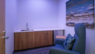 A calm, softly lit room features a comfortable chair with a decorative pillow, a countertop with a sink, and a vibrant coastal-themed artwork.