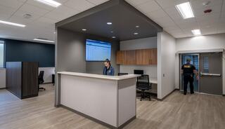 A modern reception area features a woman at a desk, a wall screen displaying information, and a man exiting through a door.