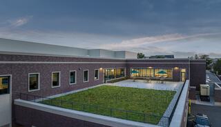 A modern building features a green rooftop and large windows, showcasing a serene outdoor area surrounded by a mix of nature and architecture.