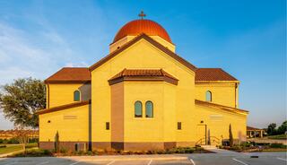 A golden-toned building features a distinctive dome with a cross, surrounded by a landscaped area and clear blue skies.