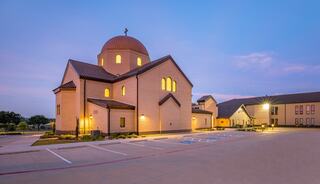 A modern church with a dome, featuring warm lights and a spacious parking area, set against a twilight sky.