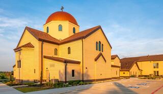 A yellow brick church features a prominent red dome and cross, surrounded by a clear sky and a spacious parking area nearby.