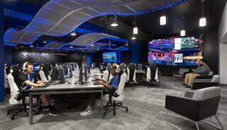 Modern esports gaming room with multiple high-tech setups, vibrant lighting, and a large screen displaying live gameplay. Relaxing seating available.