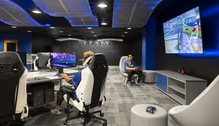 A modern gaming lounge features sleek chairs, multiple monitors, and a large screen displaying a racing game, inviting competitive play.