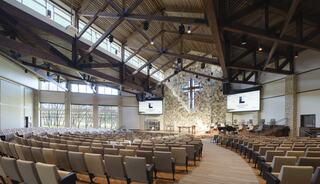 A spacious, modern worship hall featuring a wooden ceiling, a prominent cross, rows of seats, and large screens for presentations.