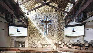 A modern church interior features a grand stone wall with a cross, flanked by large screens and arranged seating. Warm wooden details enhance the ambiance.