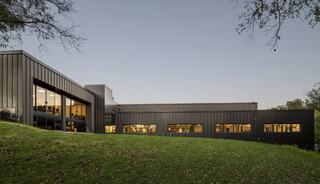 A modern building with large windows, set on a grassy slope, showcases sleek architecture against a twilight sky.