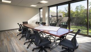 A modern conference room features a large table surrounded by black chairs and large windows offering a view of greenery outside.