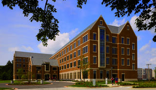 A modern, multi-story brick building with large windows, surrounded by greenery and pathways, indicative of a campus setting with people walking.