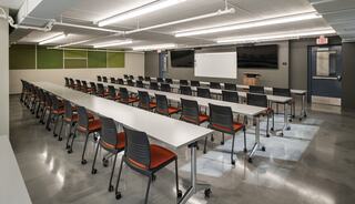 A modern classroom features rows of tables and rolling chairs, with bright lighting and a projector screen at the front.