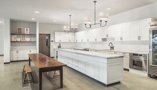 A modern kitchen features white cabinetry, sleek countertops, and a large wooden dining table, complemented by pendant lights and concrete flooring.