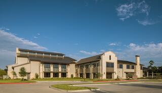 A modern building with stone accents and large windows, featuring a well-maintained exterior and surrounded by green landscaping under a blue sky.
