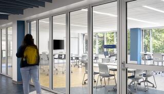 A student with a yellow backpack approaches a modern classroom with glass walls, featuring round tables and natural light.