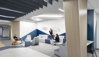 A modern, open lounge area features stylish seating in blue and gray tones, with individuals engaged in conversation and relaxing.
