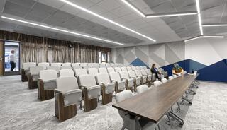 A modern classroom features rows of gray chairs, a large table, and geometric wall designs. Two people are seated in the space.
