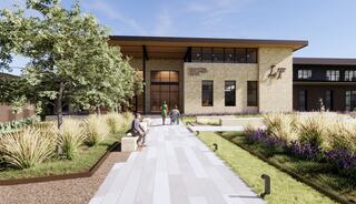 A modern building features large windows and a welcoming entrance, surrounded by landscaped gardens and walking paths, promoting community interaction.