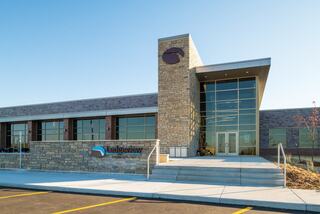 A modern building featuring a stone façade, large glass windows, and a stylish entrance, flanked by landscaped areas and a clear blue sky.