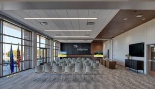 A modern meeting room features rows of chairs, large windows, a logo display, and a setup for presentations, creating a bright, inviting space.